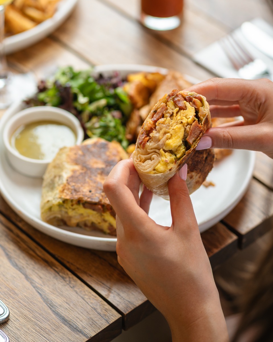 Hands holding a breakfast burrito with eggs and bacon, served with salad at Café Maud