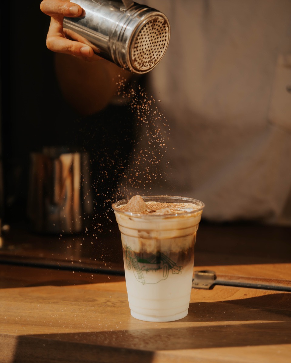 Iced coffee dusted with cocoa powder, served in a branded Café Maud cup