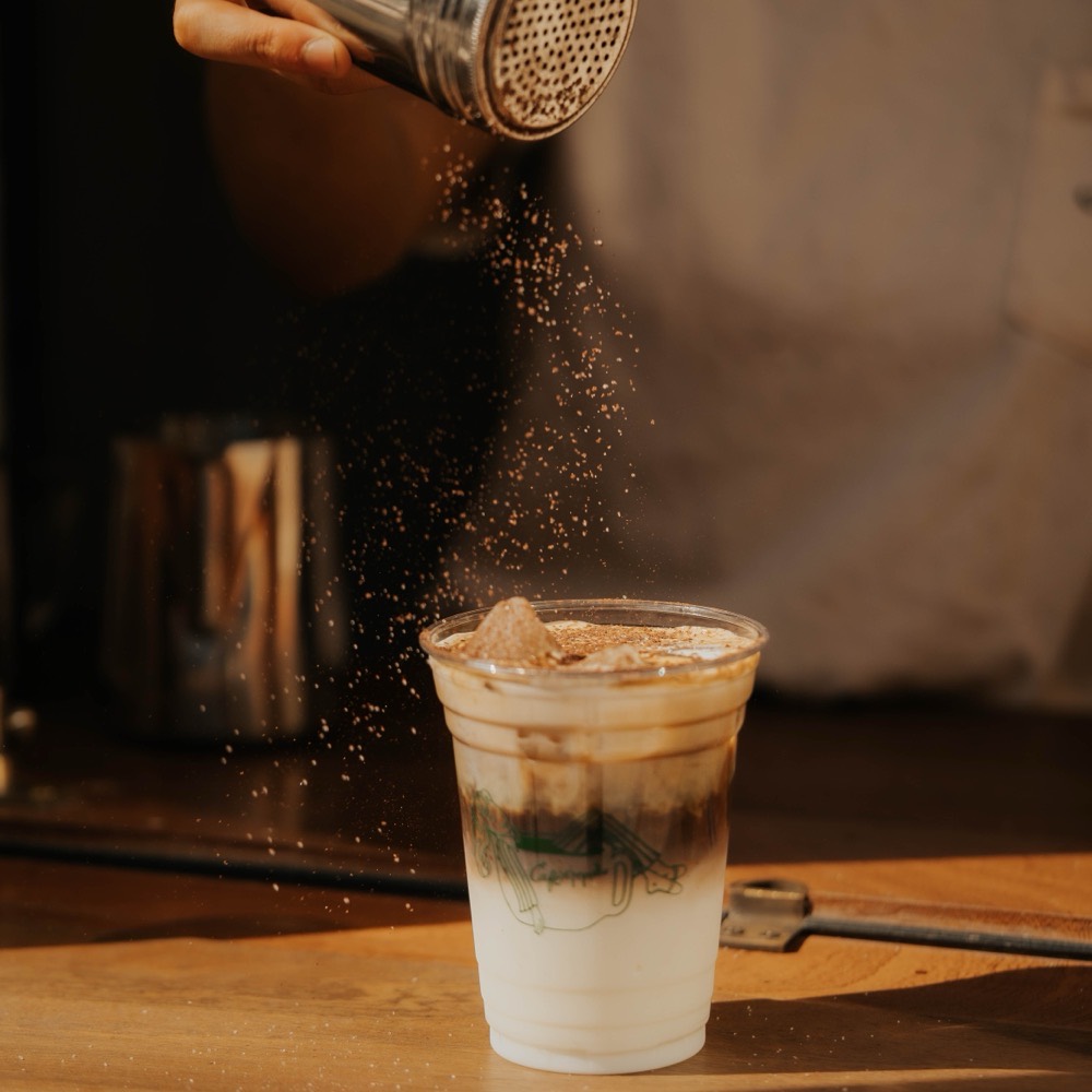 Iced coffee dusted with cocoa powder in a branded Café Maud to-go cup